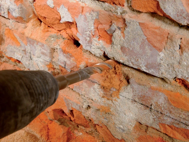 Nahaufnahme, wie mit einer Bohrmaschine Löcher in die Lagerfugen des Mauerwerks der Kellerwand gebohrt werden