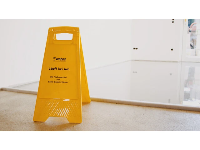 Aufnahme einer Fläche, in die eine frische Spachtelmasse eingebaut worden ist. Im Vordergrund steht ein gelber Aufsteller vor der Fläche mit der Aufschrift Läuft bei mir: Mit Fließspachtel von Saint-Gobain Weber
