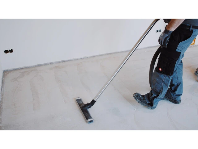Aufnahme von schräg oben, wie ein abgeschliffener Estrich mit einem Industriesauger abgesaugt wird. Rechts im Bild sieht man die Beine der Person, die den Sauger betätigt