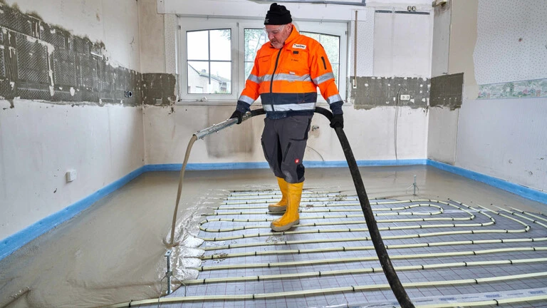 Handwerker mit oranger Arbeitsjacke, dunkelgrauer Arbeitshose, gelben Gummistiefeln und einer schwarzen Mütze auf dem Kopf steht in der Mitte eines Raumes, der gerade renoviert wird. Er steht auf den sichtbaren Rohren einer Fußbodenheizung und hat einen Schlauch in der Hand, aus dem Ausgleichsmasse auf die Rohre gepumpt wird.