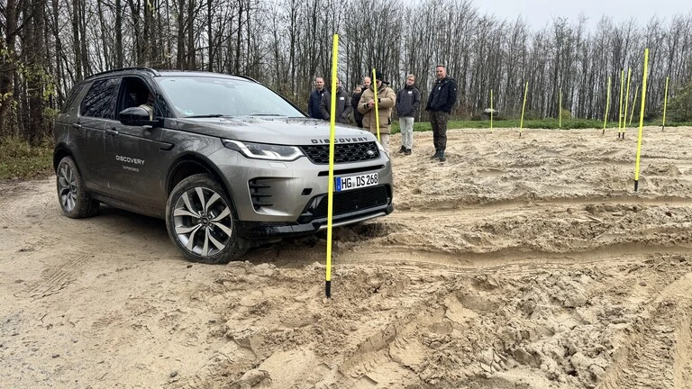 Geländewagen fährt durch einen sandigen Offroad-Parcours mit gelben Markierungsstangen. Im Hintergrund stehen mehrere Personen und beobachten das Fahrzeug