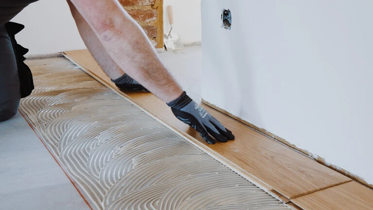 Zwei Hände drücken eine Parkett-Diele in das Klebebett des Parkettklebers ein