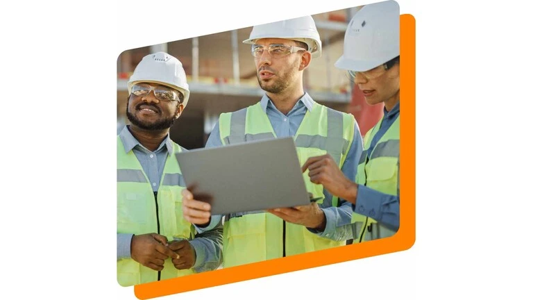 Zwei Männer und eine Frau in gelben Warnwesten und mit weißen Bauarbeiterhelmen stehen auf einer Baustelle. Der Mann in der Mitte hält einen Laptop, auf den die Frau rechts neben ihm zeigt.