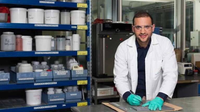 Junger Mann vor einem Materialregal mit weißem Laborkittel und grünen Handschuhen