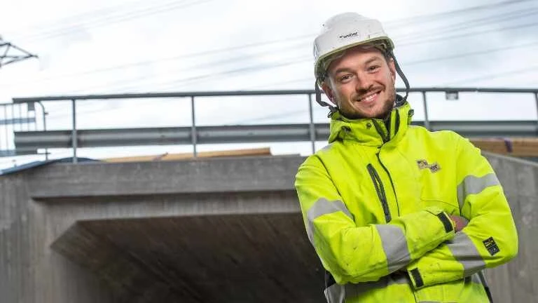 Junger Mann mit gelber Warnjacke und Baustellenhelm vor einer Betonbrücke