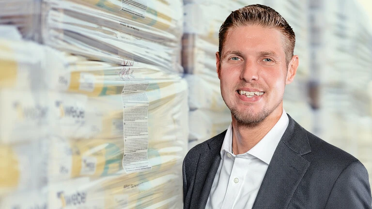 eine männliche Person im Anzug vor gestapelten, in Folie verpackten Waren in einer Lagerhalle